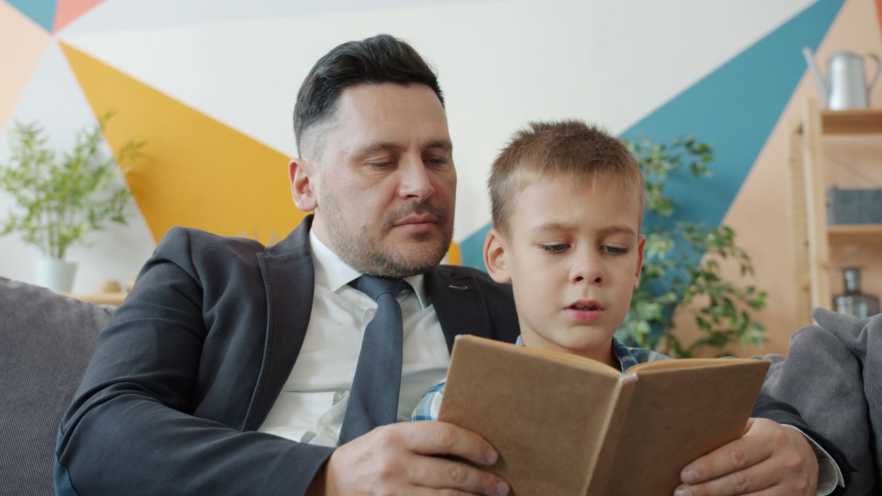 Father and son bonding over a book at home, sitting on a couch.